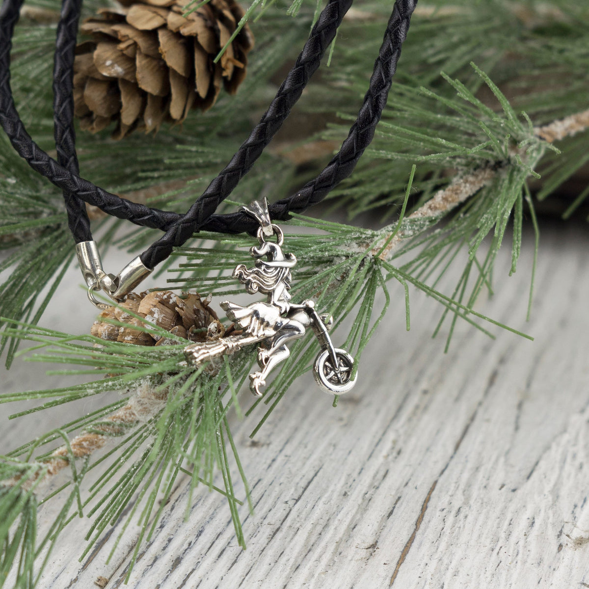 Witch on a motorcycle silver pendant on a braided black cord among pine needles
