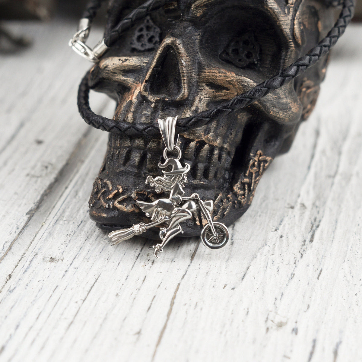 Allegra biker witch pendant in 925 sterling silver featuring a witch riding a motorcycle with a broom, displayed against a dark skull background.
