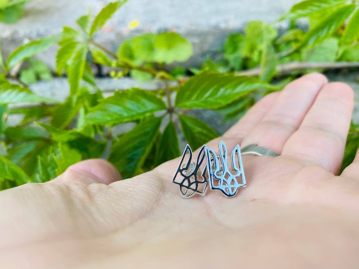 Pair of Ukrainian Tryzub stud earrings in 925 sterling silver with dark contrasting details, displayed on a hand by Yeti Jewelry