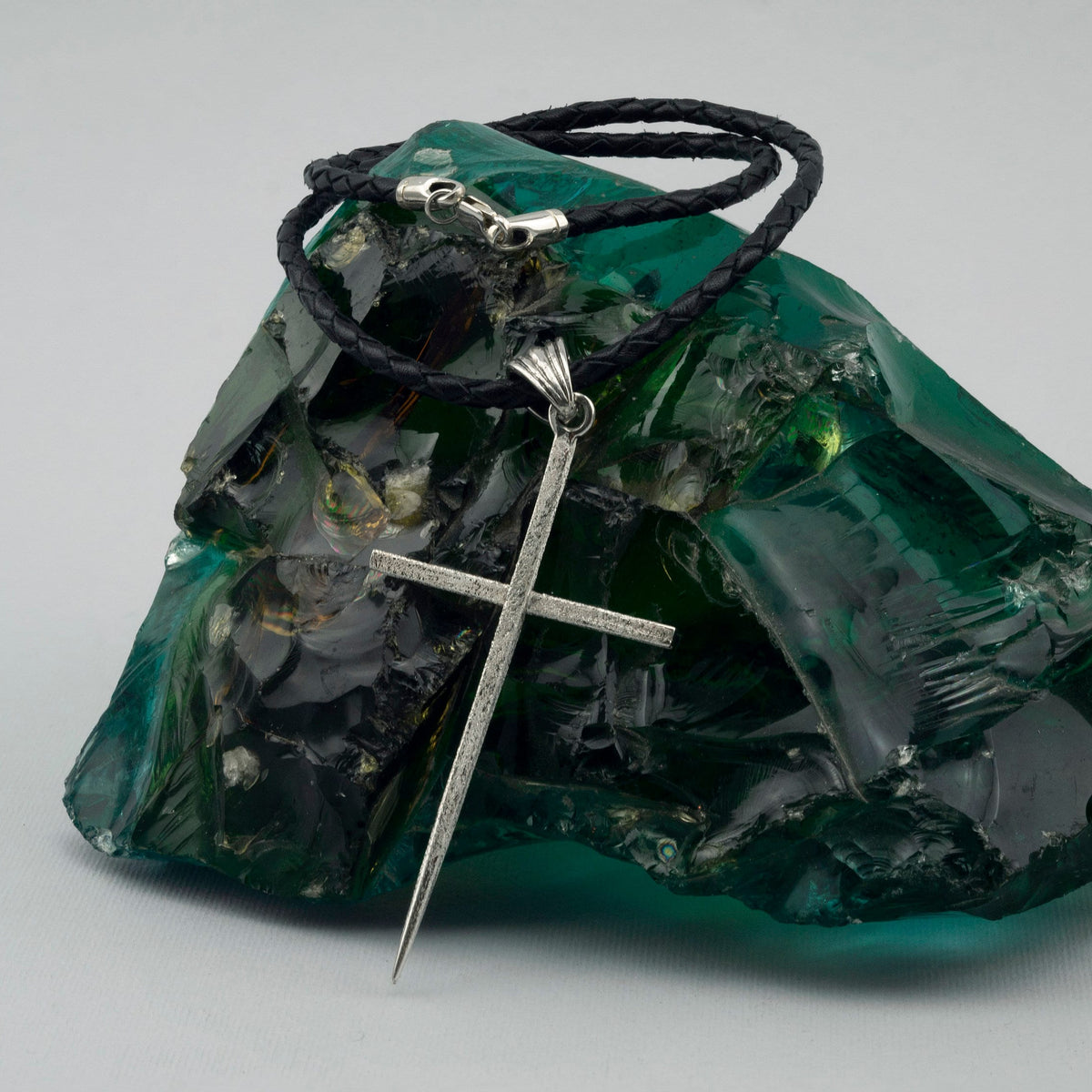 Large sterling silver spike cross pendant on a black braided leather cord, resting on green raw glass