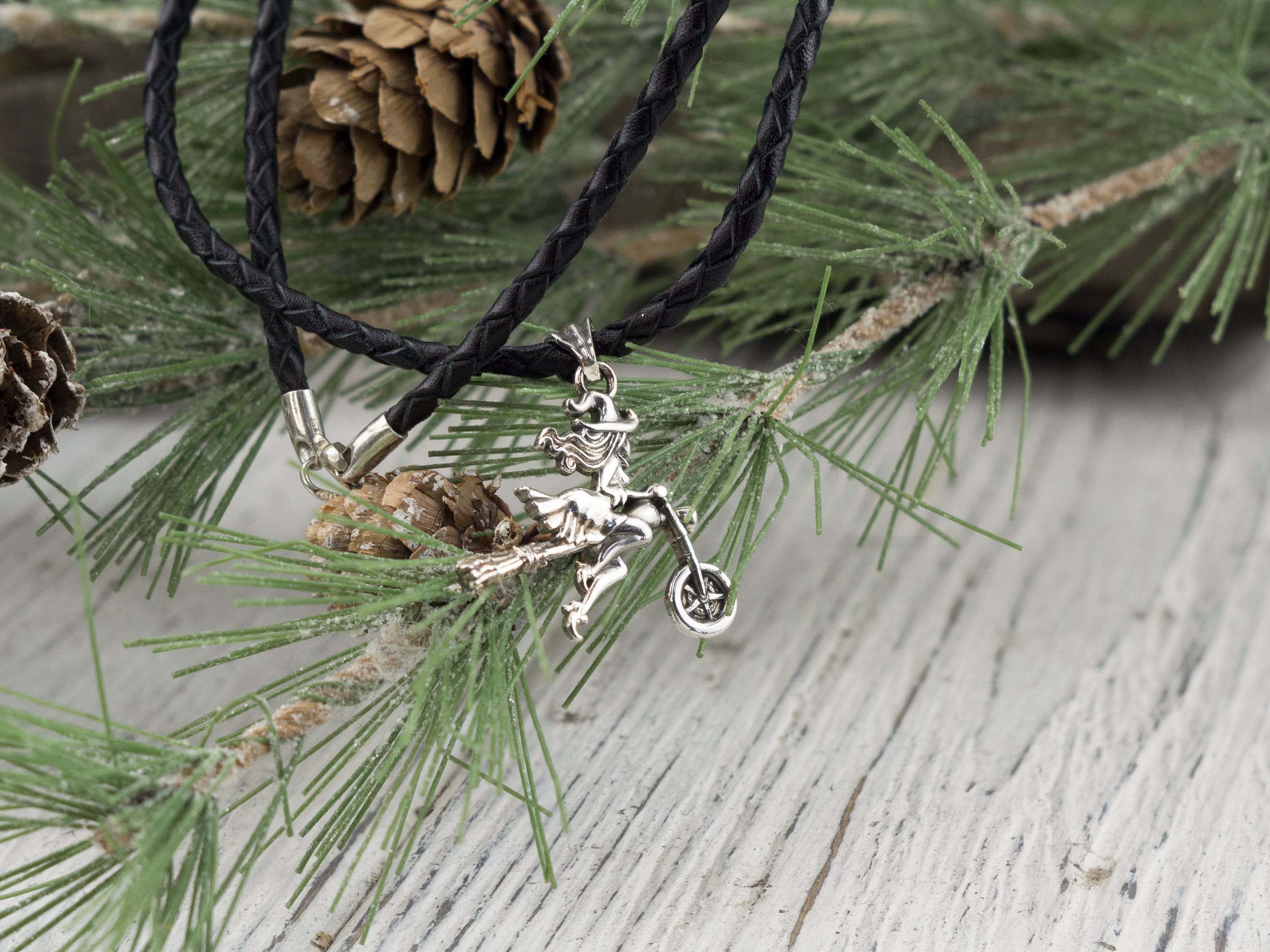Witch on a motorcycle silver pendant on a braided black cord among pine needles