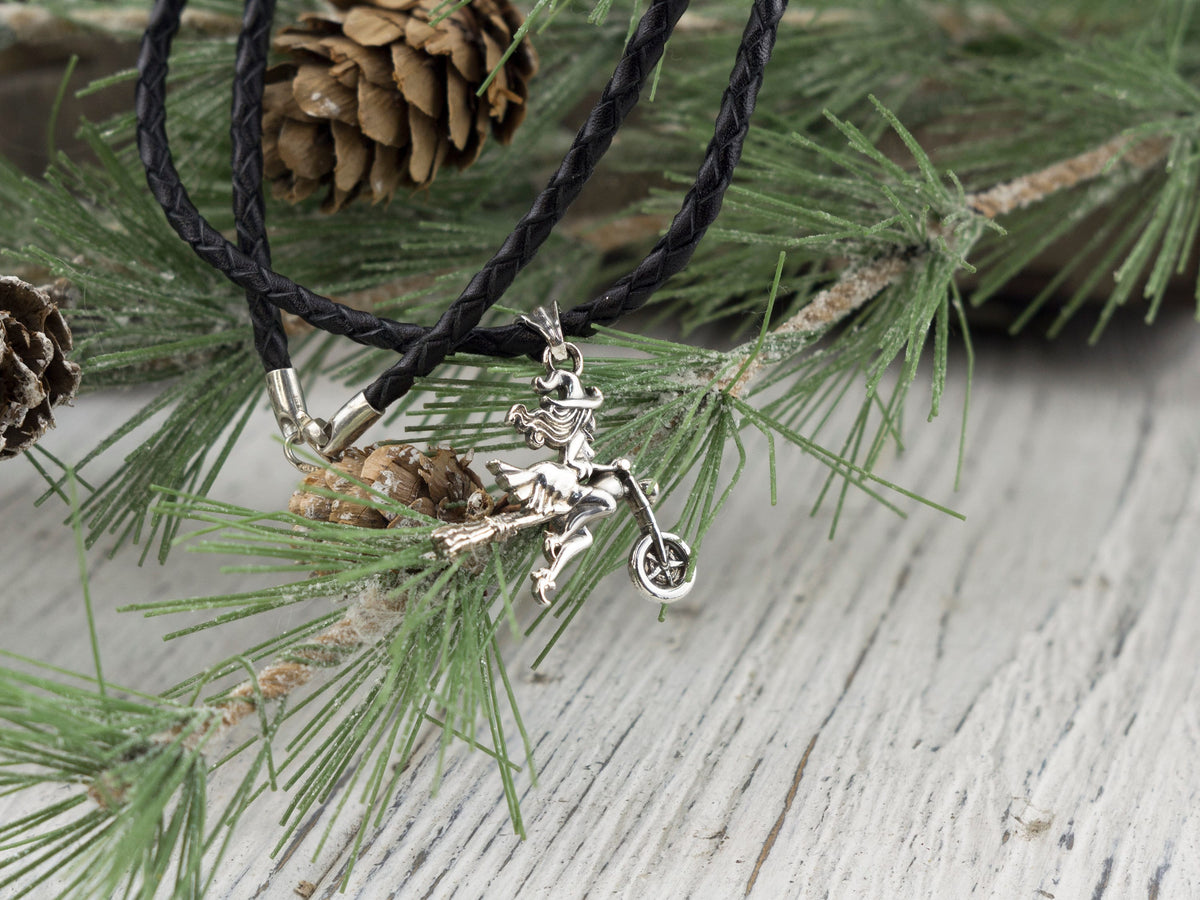 Witch on a motorcycle silver pendant on a braided black cord among pine needles
