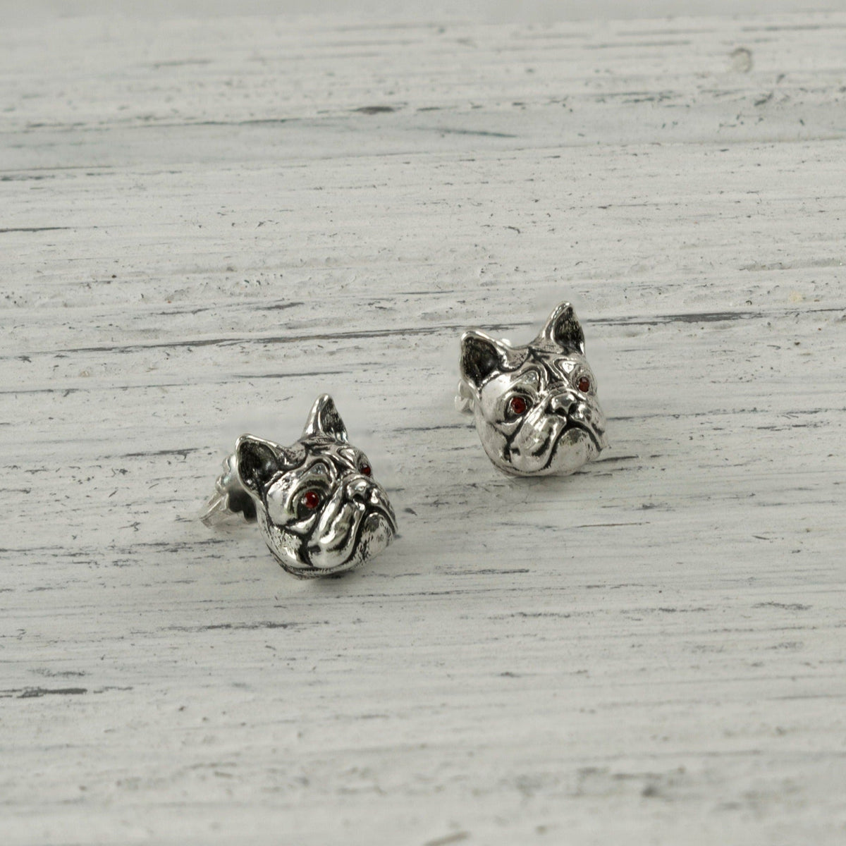 Pair of silver French bulldog stud earrings on white wood, angled view