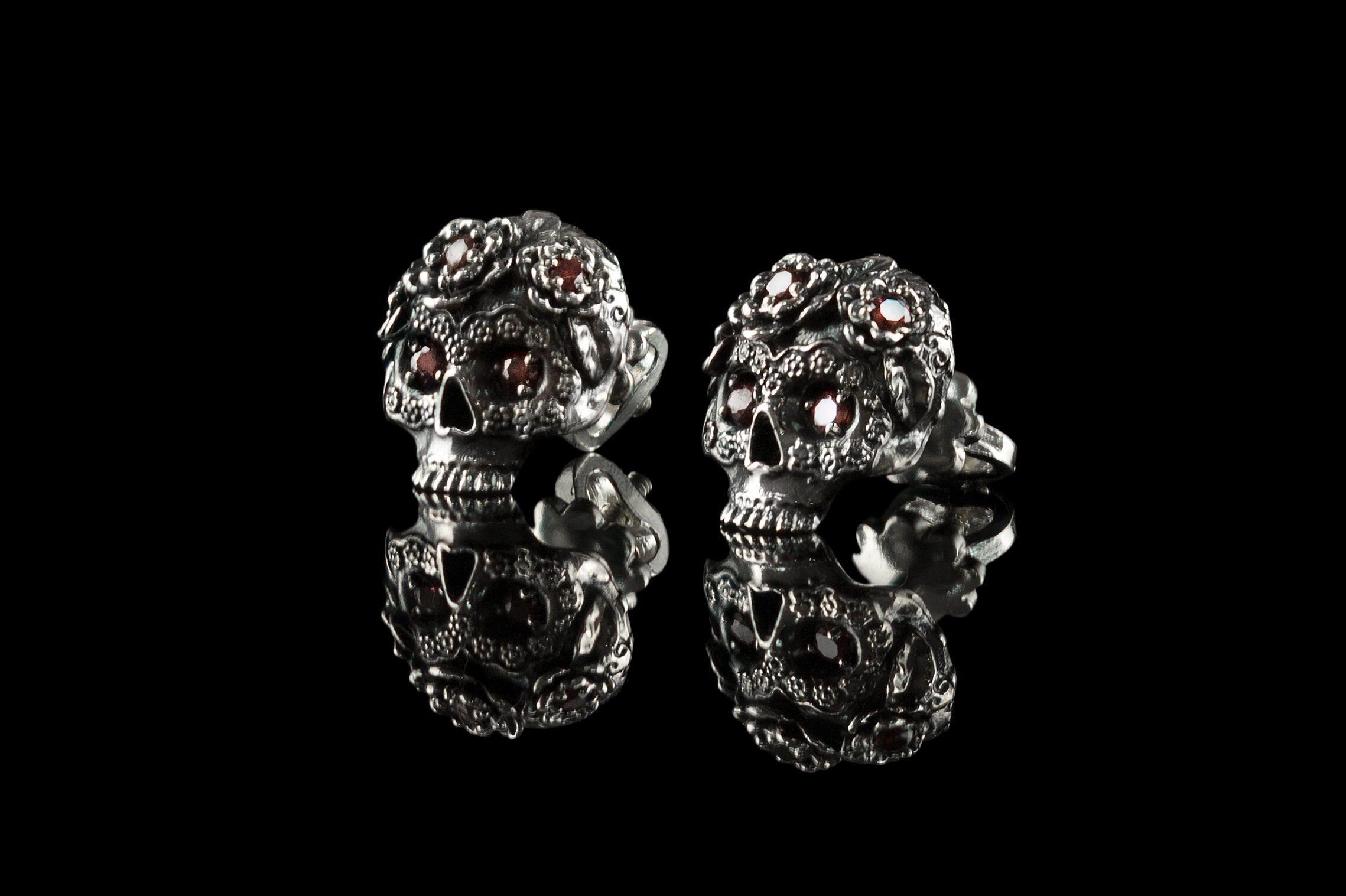 Pair of Frida skull stud earrings with red CZ and reflection, angled view