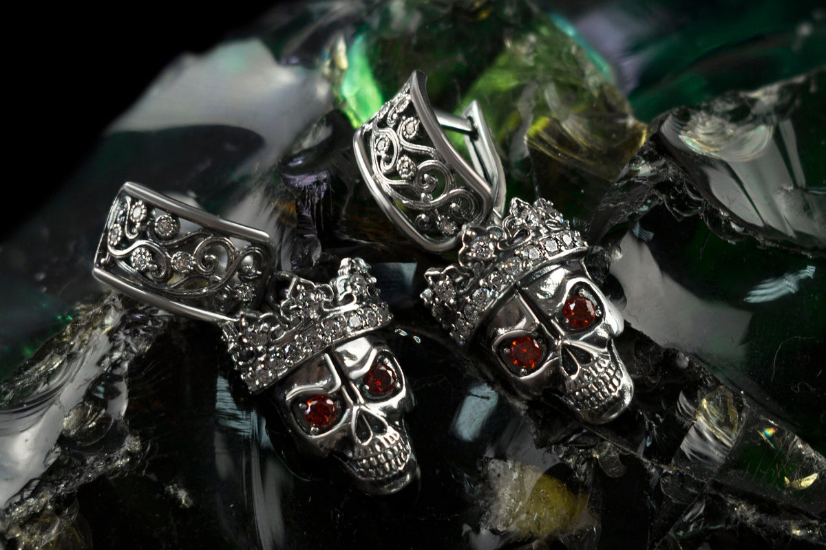 Close-up of Santa Muerte-inspired sterling silver skull earrings showing detailed crowns and red cubic zirconia eyes