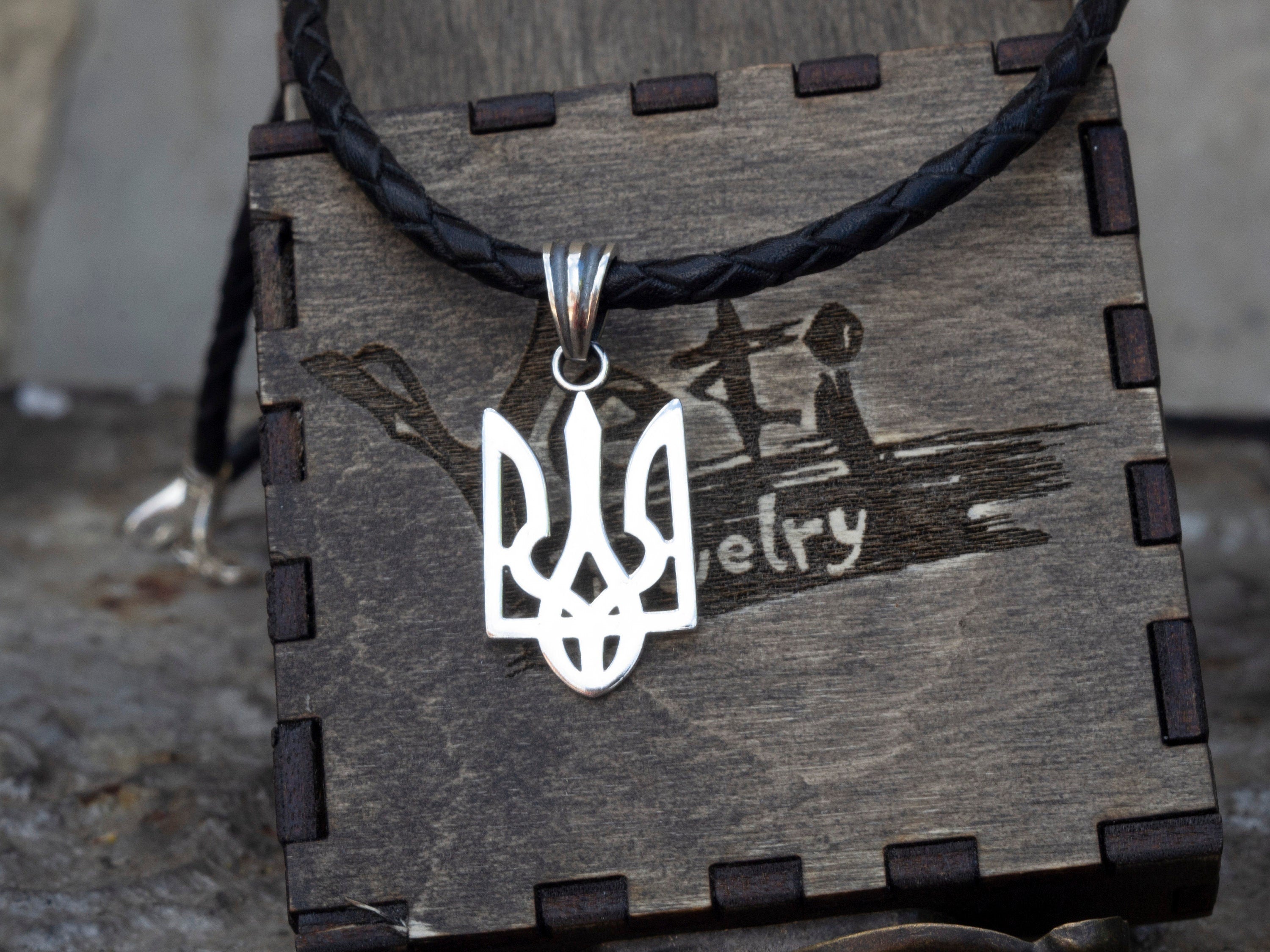 Ukrainian Tryzub pendant in polished 925 sterling silver displayed on a black braided leather cord against a branded wooden jewelry box.