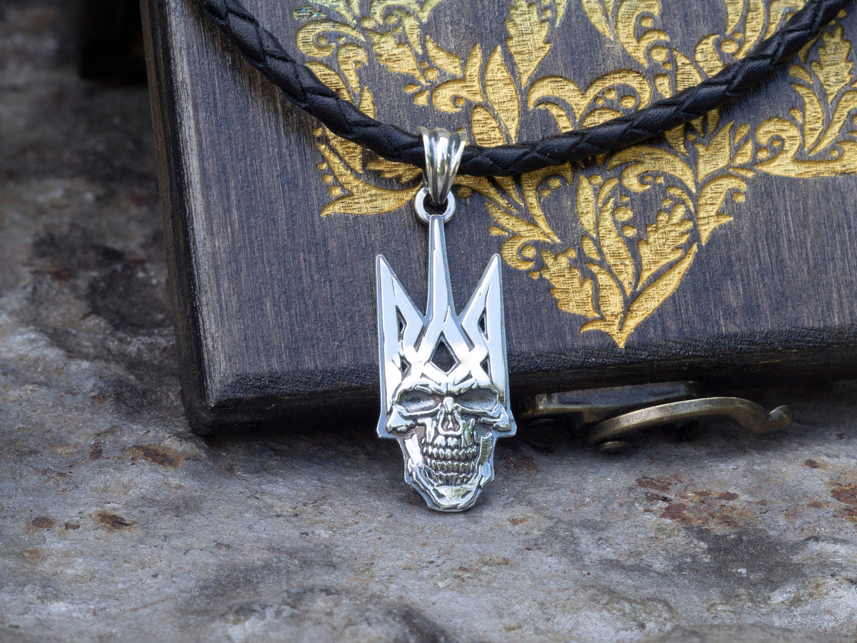Handcrafted 925 sterling silver Ukrainian Tryzub pendant integrated with a sculpted skull, hanging on a black braided leather cord against a branded wooden box