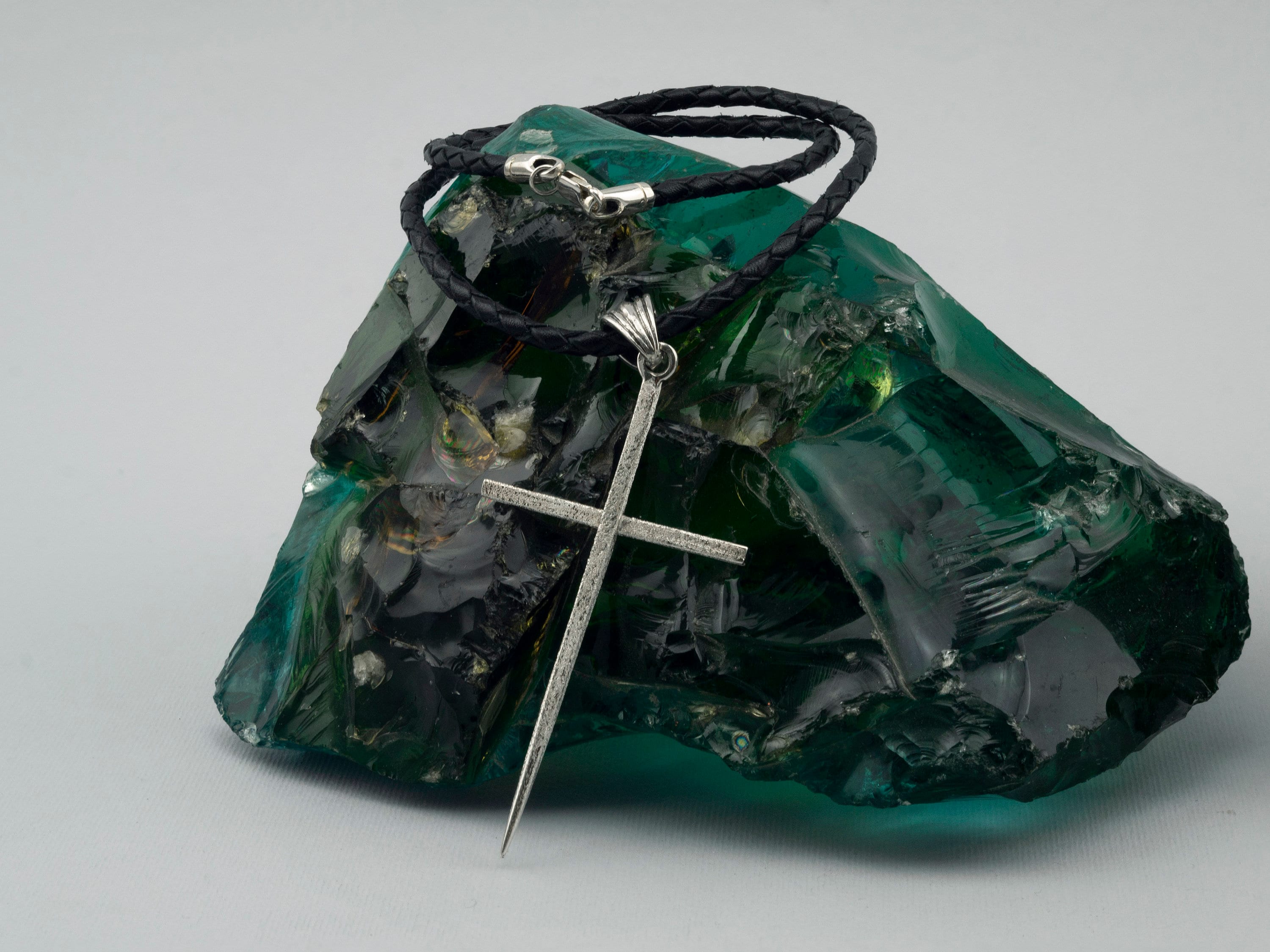 Large sterling silver spike cross pendant on a black braided leather cord, resting on green raw glass