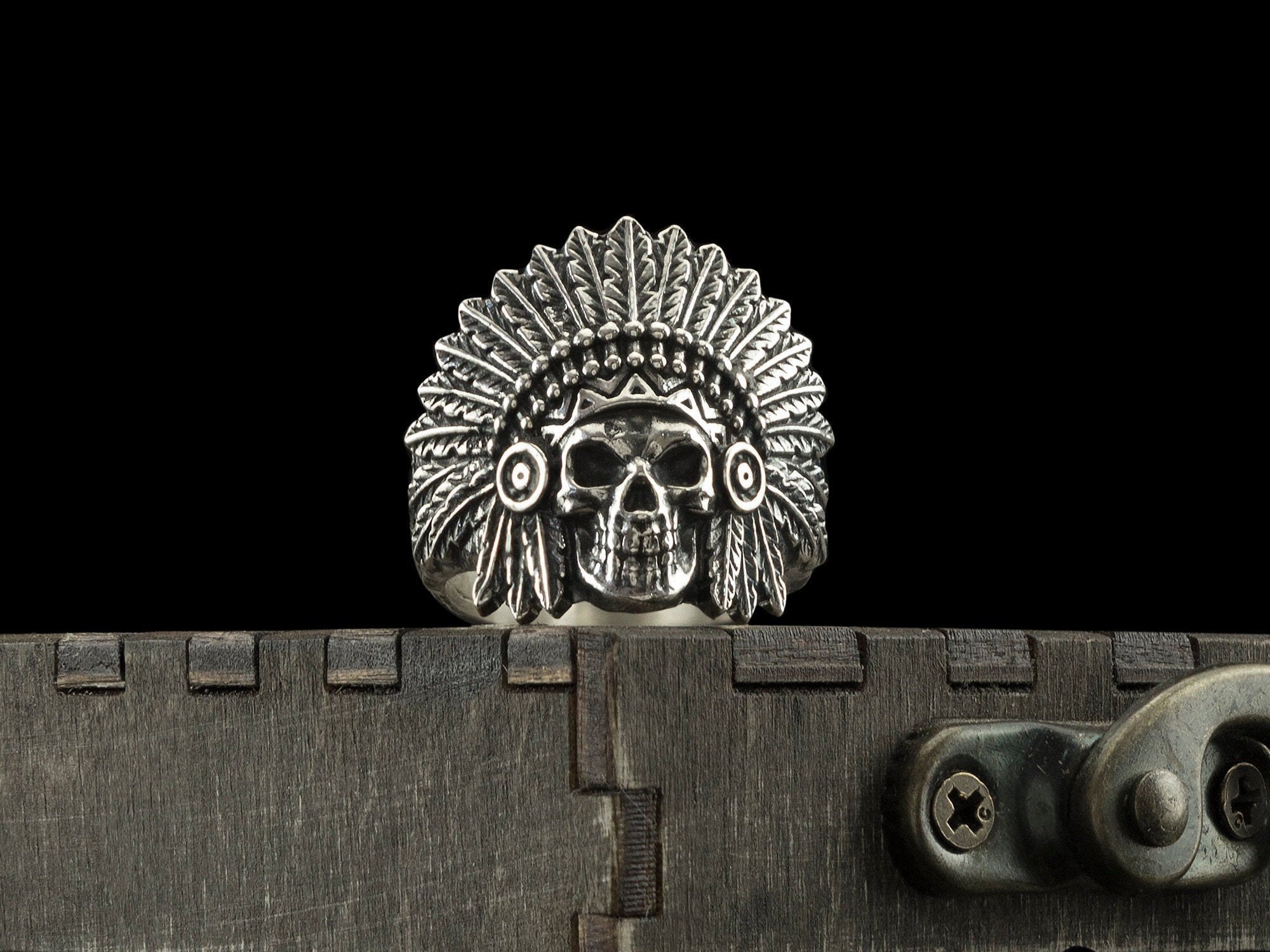 War Bonnet chief skull ring in sterling silver, front view of the feathered headdress on a wooden box